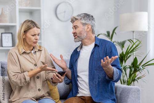 A couple seated on a couch engaged in an argument over a smartphone, depicting emotions of jealousy and conflict possibly due to social media.