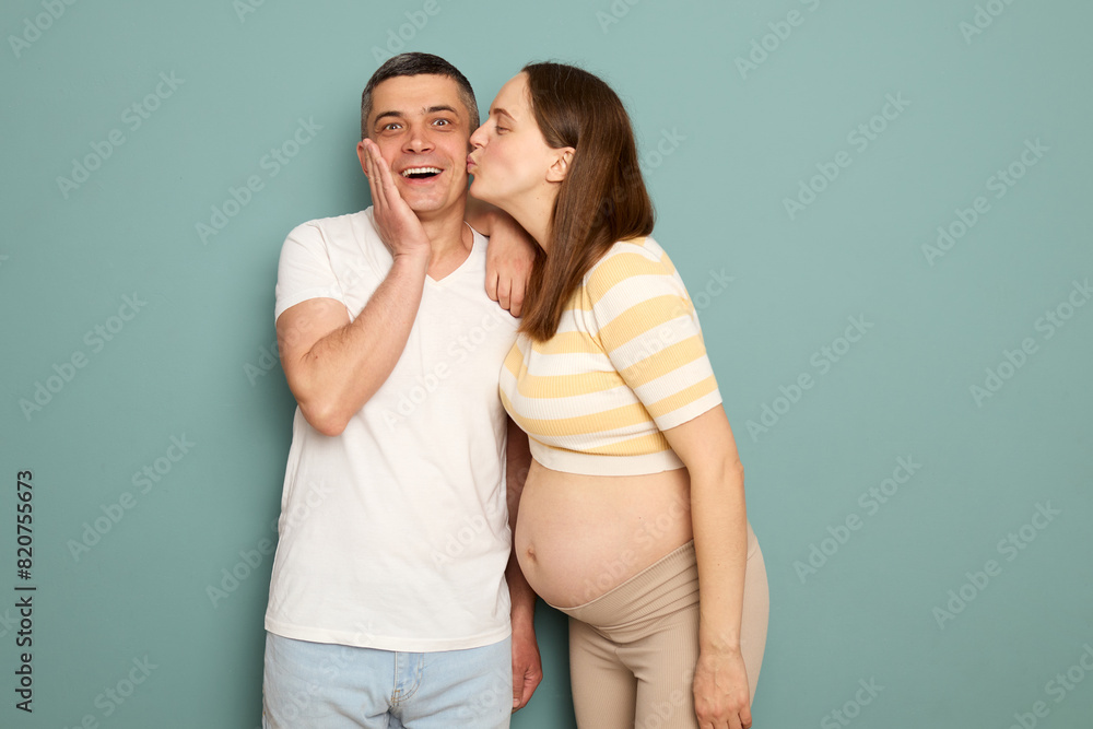 Naklejka premium Happy cheerful young couple pregnant woman and man standing isolated over light green background wife kissing surprised husband cheek with love
