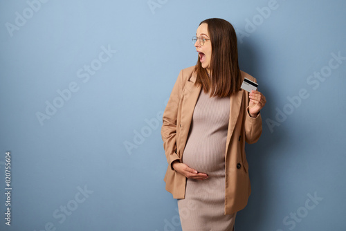 Surprised pregnant woman wearing dress and jacket isolated over blue background looking away at advertisement area screaming with excitement looking at free space for promotion