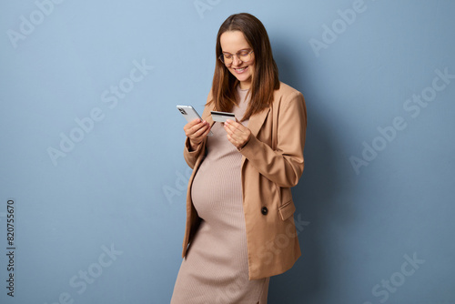 Smiling happy pregnant woman wearing dress and jacket isolated over blue background making online payment with smartphone and credit card enjoying online shopping entering bank data