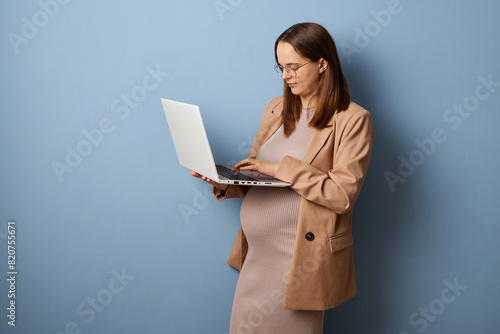 Confident brown haired pregnant woman wearing dress and jacket isolated over blue background using laptop for remote job typing on keyboard doing internet project
