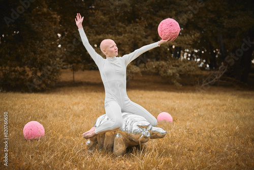 Young hairless girl with alopecia in white cloth sits on tardigrade figure and holds pink ball in hand on fall lawn park, surreal scene with bald teenage girl engages with symbolic elements photo