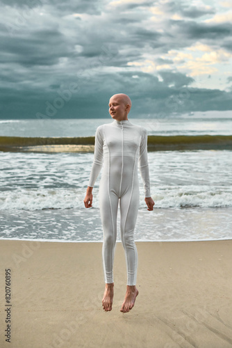 Candid hairless girl with alopecia in white futuristic suit twirls in jump on sea sandy beach, metaphoric scene with bald teenage girl creates whirlwind of positive energy and confidence © TRAVELARIUM