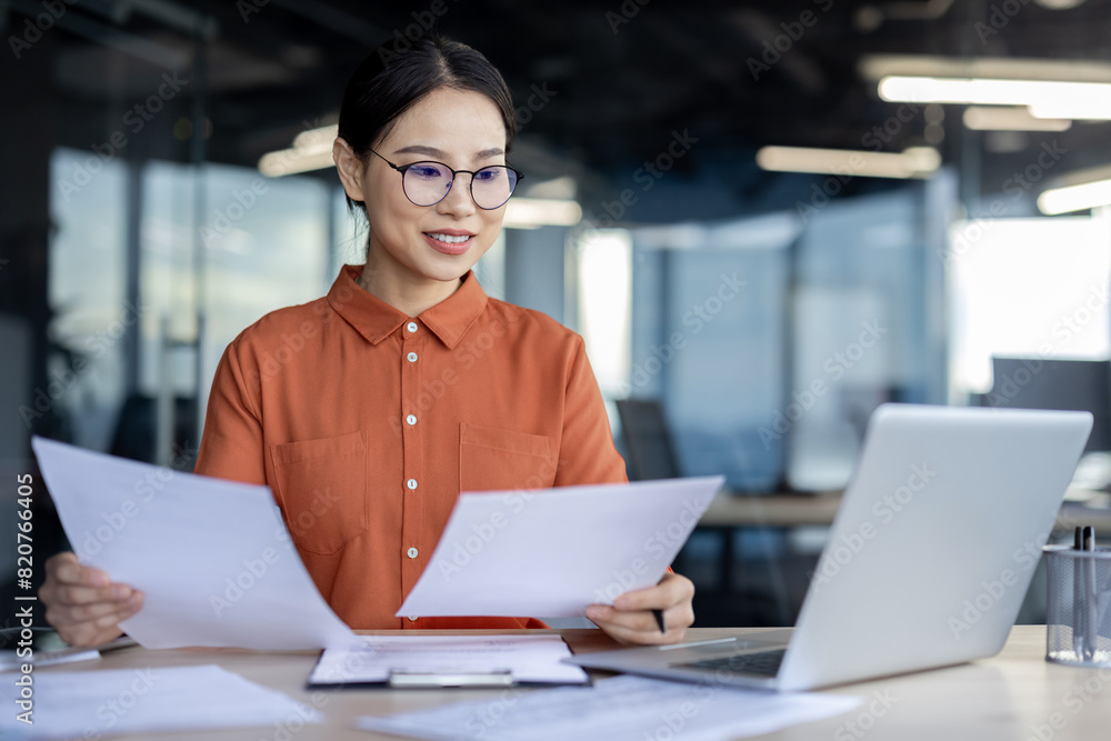 Asian businesswoman analyzing documents at desk in a modern office while working on a laptop. Professional, focused, and productive work environment. Asian businesswoman analyzing documents at desk in a modern office while working on a laptop. Professional, focused, and productive work environment.
