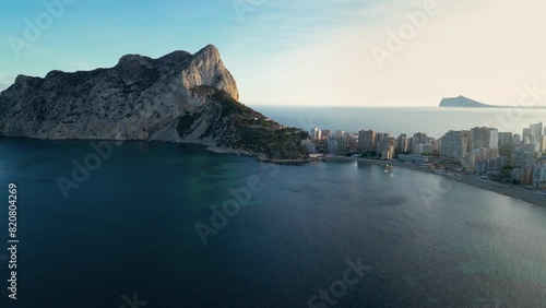 Costal city Calpe at sunset. View of part of the city and famous Peñón de Ifach Mountain and Natural Park. Sun reflection on mountain and buildings. Travel destination. Drone slowly ascending. photo