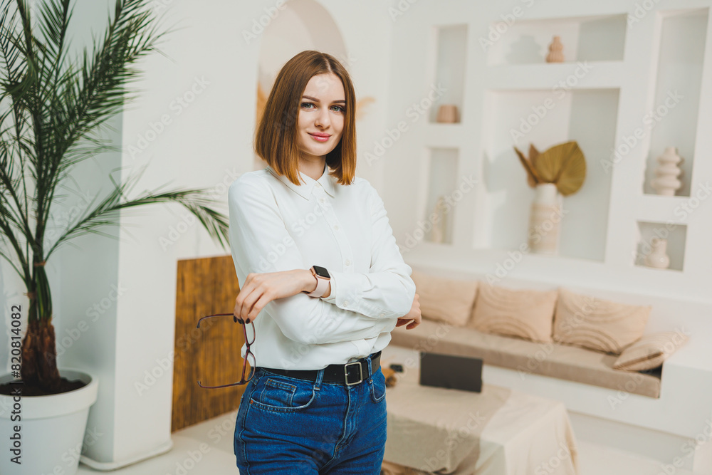 A pretty woman with glasses is standing in the middle of a bright living room, wearing a white t-shirt and jeans. Young confident woman working remotely.