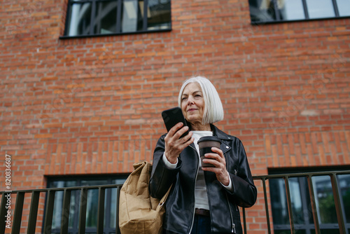 Portrait of stylish mature woman with gray hair on city street, holding smartphone and travel mug with coffee. Older woman in leather jacket with soft smile.
