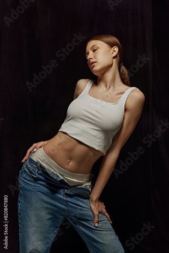 Beautiful young girl with slim body and nude makeup, wearing white top and jeans, posing against black background. Concept of beauty, body, sport and health, fashion, wellness
