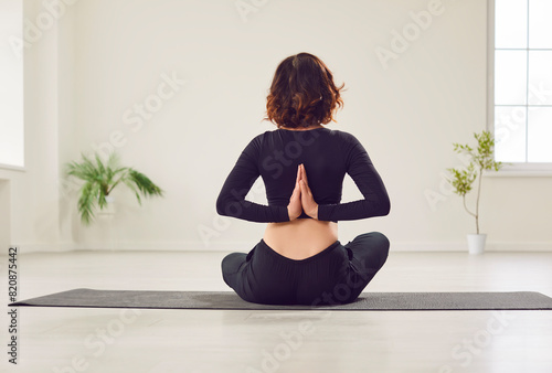 Yogi woman rear view practicing yoga, sitting in Ardha Padmasana exercise, Half Lotus pose with namaste gesture behind her back, working out on mat, wearing balck sportswear, in white studio, home photo