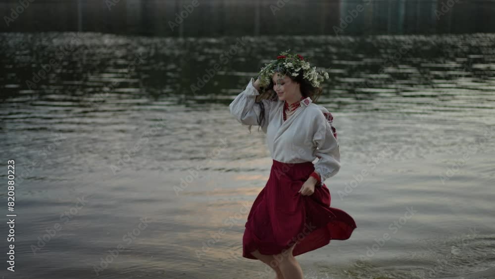 Slow motion. A girl in a traditional Ukrainian costume and with a wreath of wild herbs and flowers on her head runs barefoot through the water along the sandy bank of the river. The girl holds the hem Slow motion. A girl in a traditional Ukrainian costume and with a wreath of wild herbs and flowers on her head runs barefoot through the water along the sandy bank of the river. The girl holds the hem