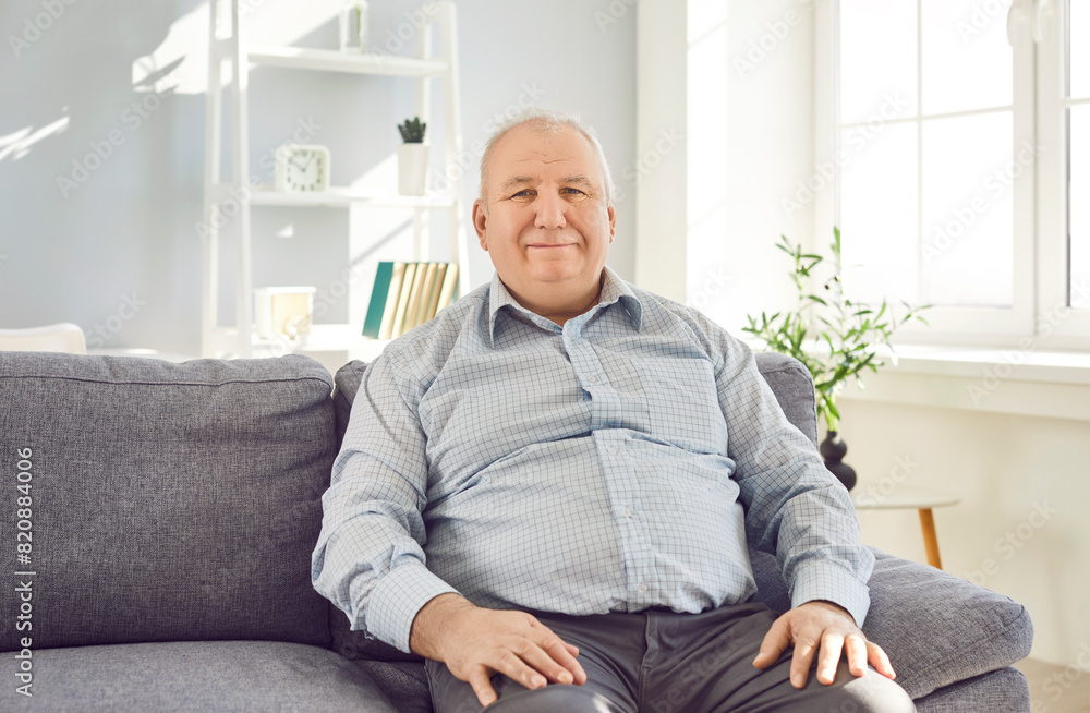Fototapeta premium Portrait of confident smiling elderly retired man sitting on sofa in living room at home and looking cheerful at camera. Gray-haired senior man wearing shirt resting on couch enjoying retirement.