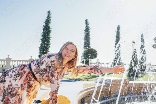 Young cheerful woman in light summer dress with floral pattern touches jet of water in fountain.