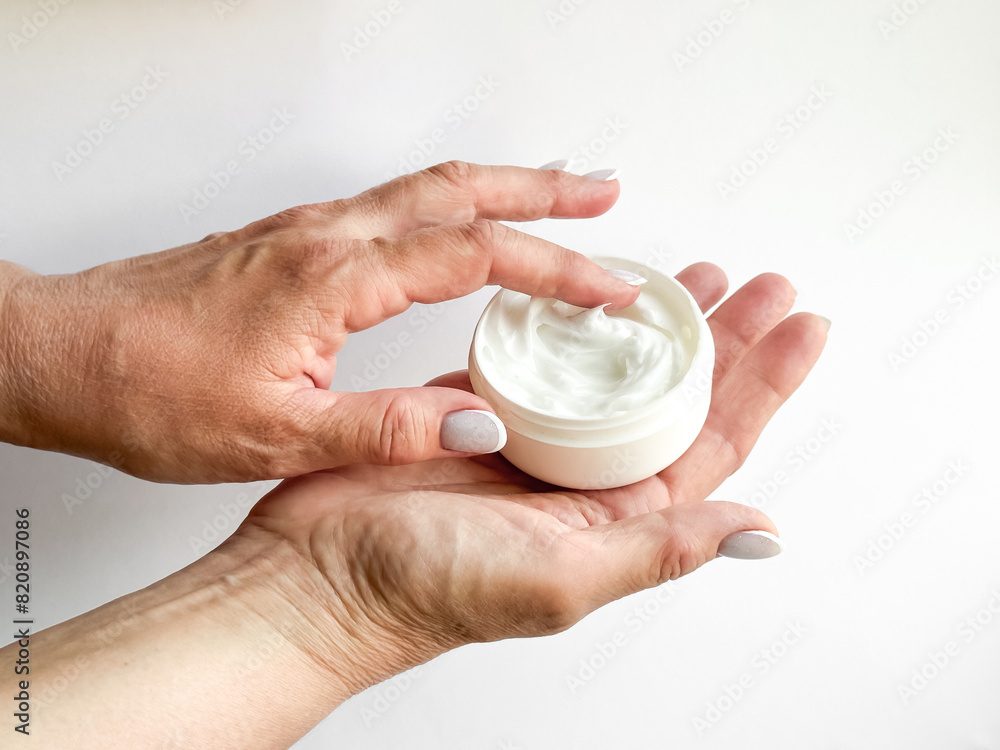 Close up of middle age woman hands holding container of cream and dipping finger on white background. Skincare and moisturizing concept. For healthcare, wellness, and self care poster. Close up of middle age woman hands holding container of cream and dipping finger on white background. Skincare and moisturizing concept. For healthcare, wellness, and self care poster.