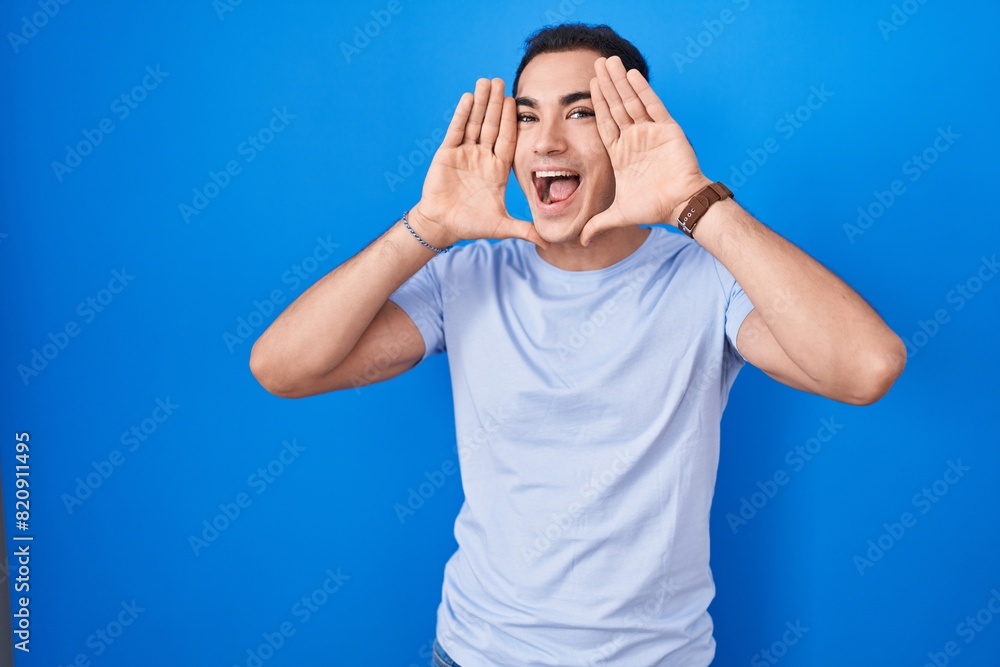 Young hispanic man standing over blue background smiling cheerful playing peek a boo with hands showing face. surprised and exited Young hispanic man standing over blue background smiling cheerful playing peek a boo with hands showing face. surprised and exited