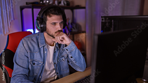 Concentrated young man with a beard wearing headphones in a dark gaming room looking at a computer screen.