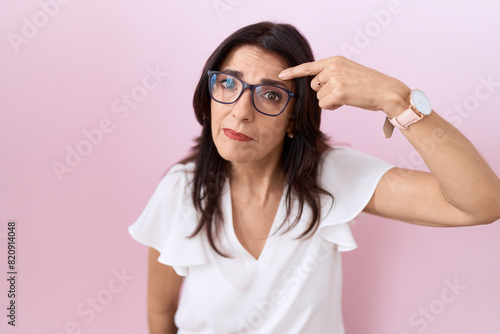 Middle age hispanic woman wearing casual white t shirt and glasses pointing unhappy to pimple on forehead, ugly infection of blackhead. acne and skin problem
