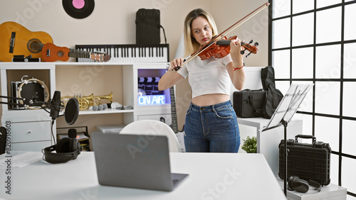 Young blonde woman musician having online violin lesson by laptop at music studio