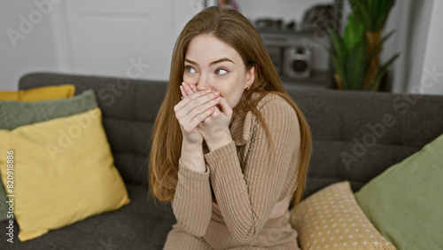 A surprised young woman sitting on a sofa at home, covering her mouth with hands, looking away.
