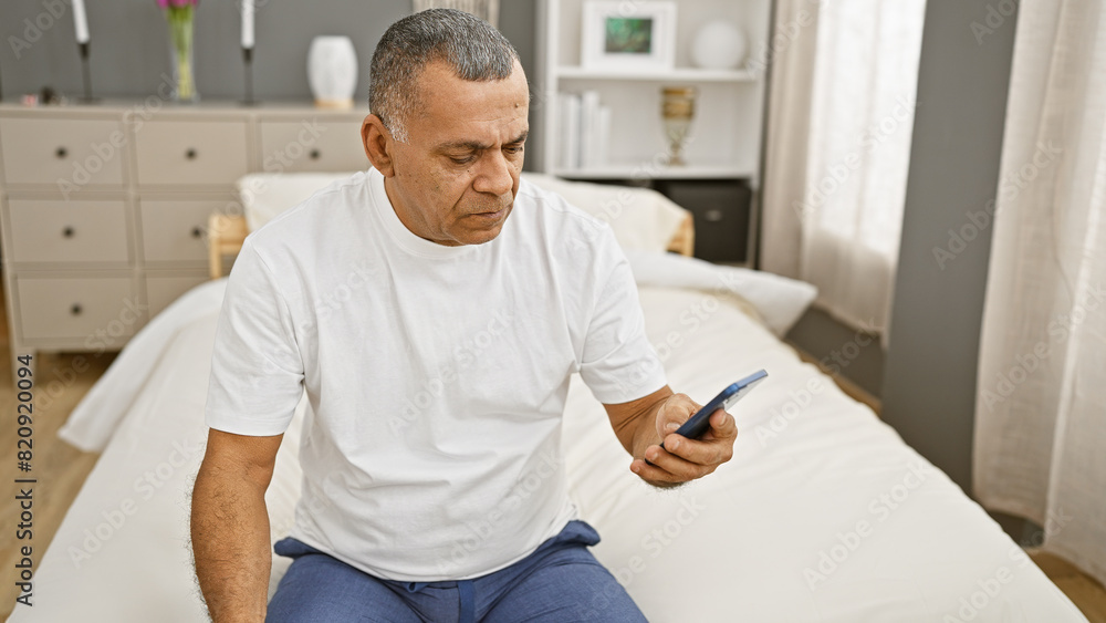 Mature hispanic man sitting on bed in bedroom looking at smartphone indoors. Mature hispanic man sitting on bed in bedroom looking at smartphone indoors.