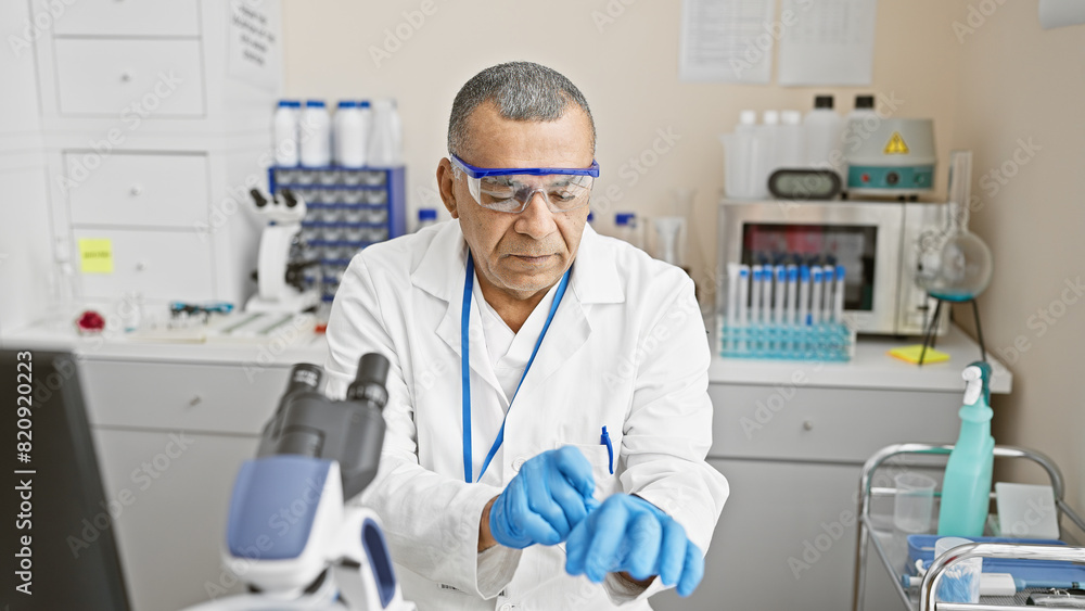 Mature hispanic man examines samples in a medical laboratory indoor setting wearing safety glasses and gloves. Mature hispanic man examines samples in a medical laboratory indoor setting wearing safety glasses and gloves.