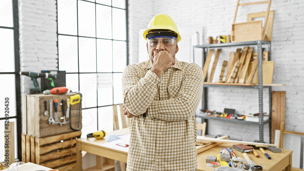 Pensive hispanic man in hardhat standing indoors at a carpentry workshop surrounded by tools and wood. Pensive hispanic man in hardhat standing indoors at a carpentry workshop surrounded by tools and wood.