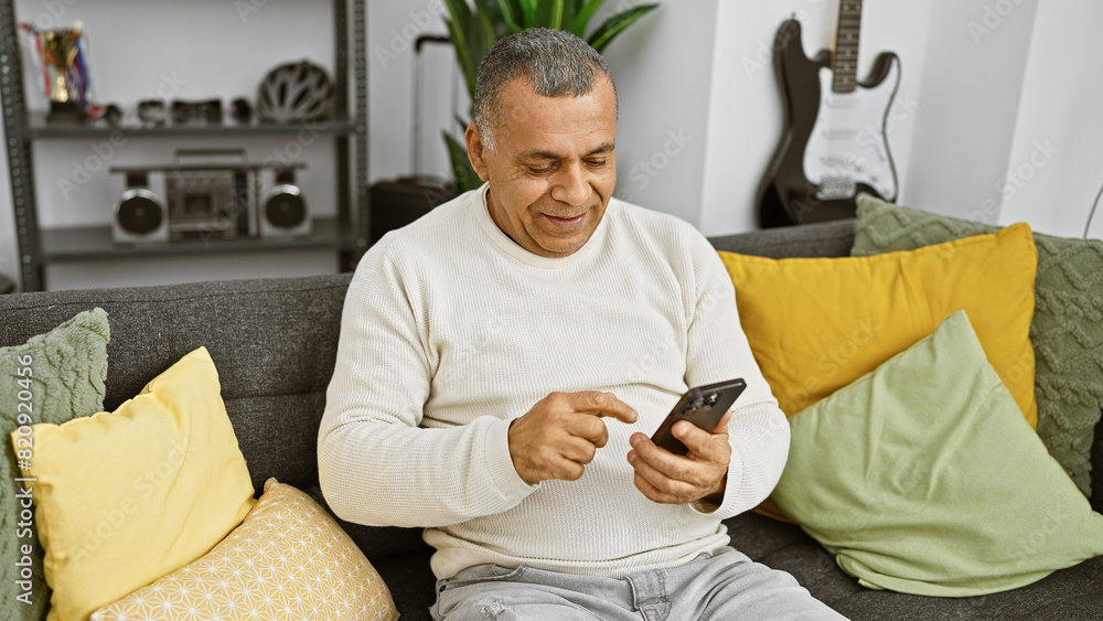 Obraz premium Smiling middle-aged hispanic man using a smartphone in a cozy modern living room
