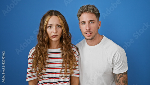 A couple stands together against a blue background, their expressions serious and connected.