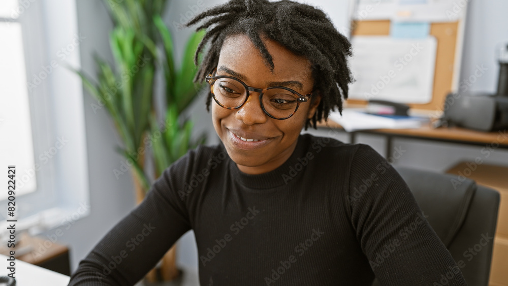 A smiling young black woman with dreadlocks wearing glasses, seated indoors at an office workplace. A smiling young black woman with dreadlocks wearing glasses, seated indoors at an office workplace.