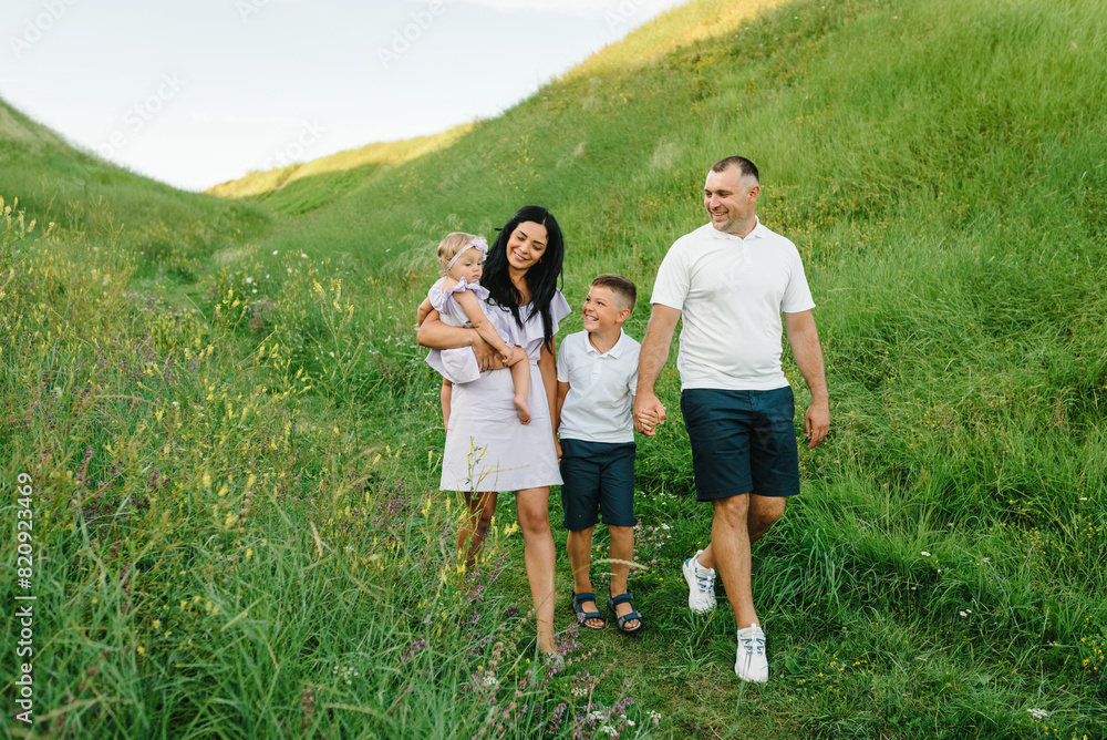 Children hold hands parents in nature. Mom, dad, daughter girl and son boy walking in green grass in field. Happy young family spending time together outdoors. Family holiday at sunset on summer day. Children hold hands parents in nature. Mom, dad, daughter girl and son boy walking in green grass in field. Happy young family spending time together outdoors. Family holiday at sunset on summer day.