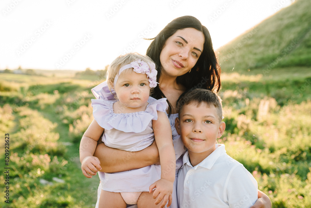 Mom hugs children standing in field at sunset. Portrait of daughter toddler and son hugging mother in nature on summer day. Concept of friendly family. Closeup. Mommy and kids spend time together. Mom hugs children standing in field at sunset. Portrait of daughter toddler and son hugging mother in nature on summer day. Concept of friendly family. Closeup. Mommy and kids spend time together.