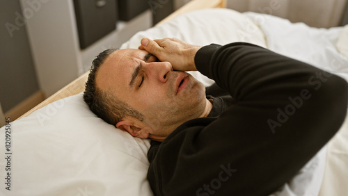 A young hispanic man appears distressed on a bed in a bedroom, indoors.