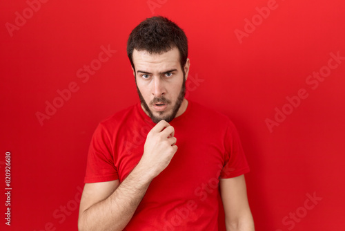Young hispanic man wearing casual red t shirt looking fascinated with disbelief, surprise and amazed expression with hands on chin
