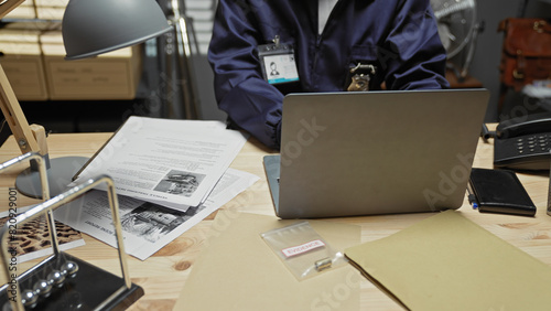 An african american male detective analyzes evidence in a police station office with a laptop and documents on the desk.