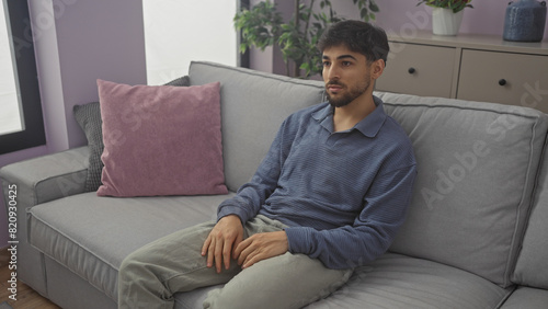 Young, bearded man sitting thoughtfully on a grey sofa in a modern living room, portraying comfort and relaxation at home. photo