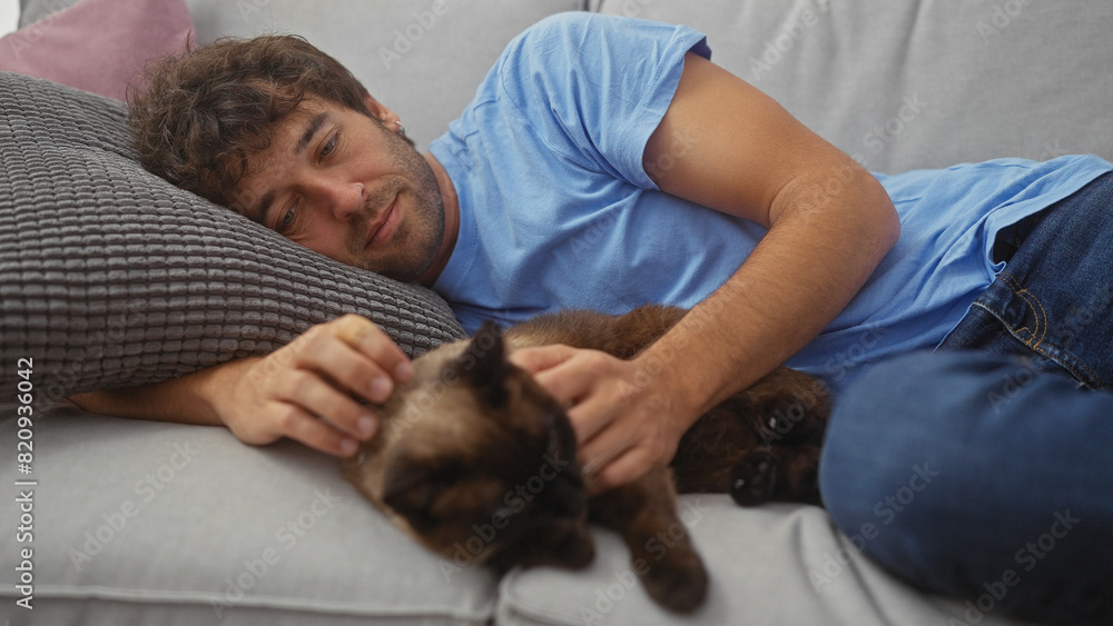 Young hispanic man relaxing on couch with his pet siamese cat in a cozy home interior. Young hispanic man relaxing on couch with his pet siamese cat in a cozy home interior.