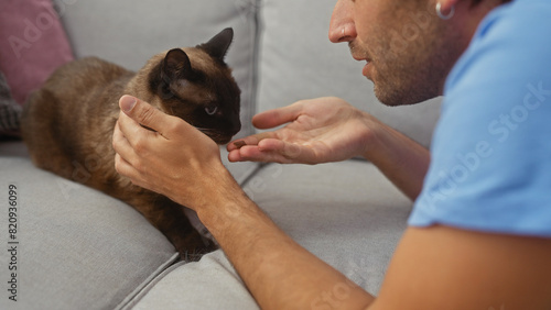 A young man at home gently interacts with his siamese cat on a cozy sofa  exemplifying a serene indoor moment.