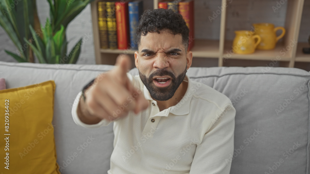 Angry young hispanic man with beard pointing at camera in a cozy living room setting Angry young hispanic man with beard pointing at camera in a cozy living room setting