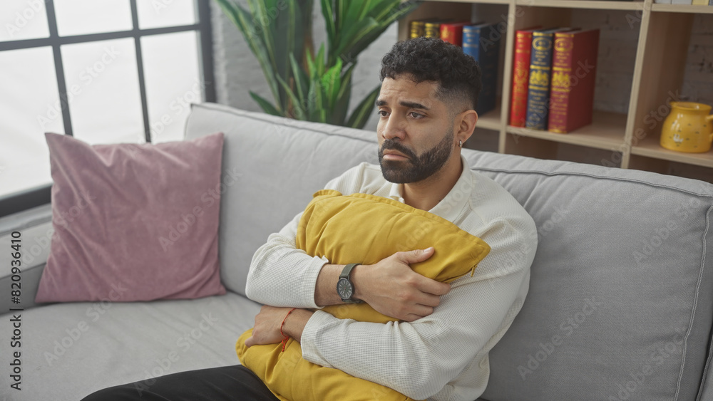 A pensive young hispanic man with a beard hugs a yellow cushion sitting on a gray couch in a modern living room. A pensive young hispanic man with a beard hugs a yellow cushion sitting on a gray couch in a modern living room.