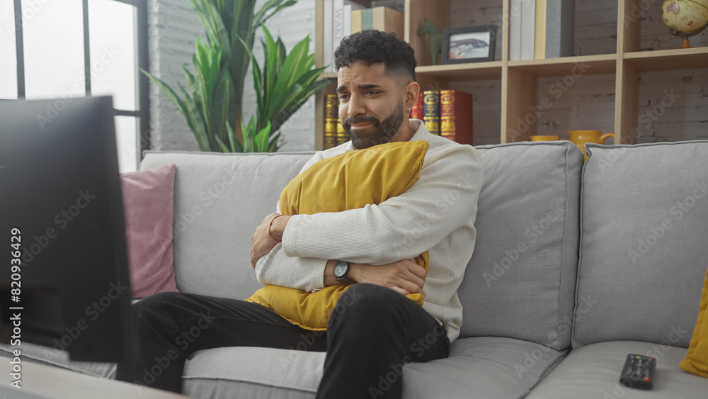 Handsome hispanic man with beard sitting on a gray sofa looking sad while watching tv in a cozy living room. Handsome hispanic man with beard sitting on a gray sofa looking sad while watching tv in a cozy living room.
