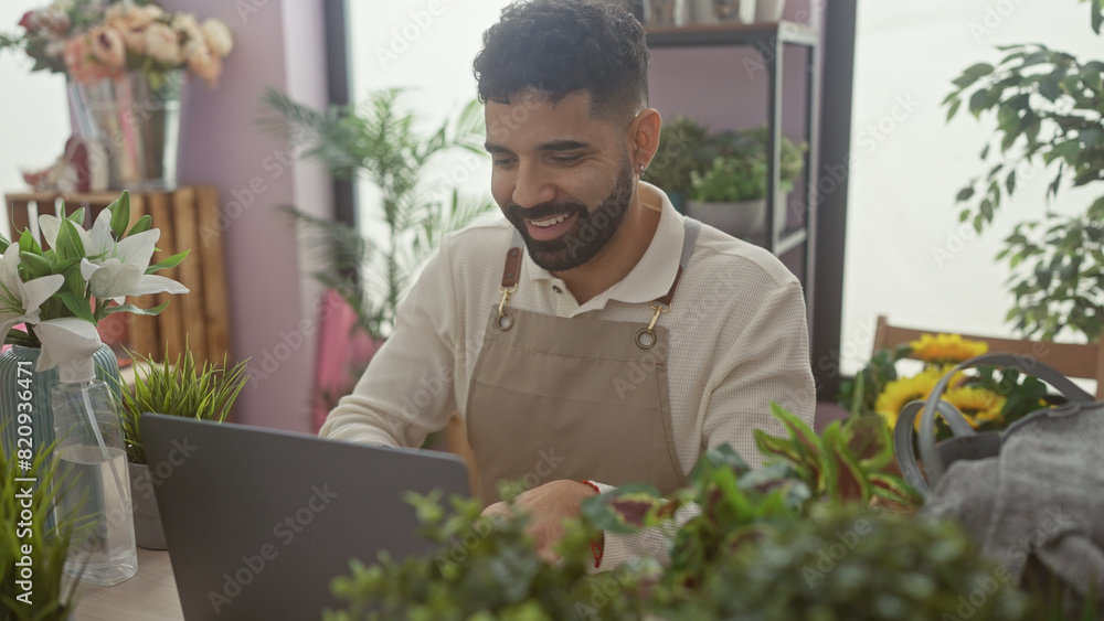 A cheerful hispanic man with a beard works on a laptop in a flower shop filled with various plants and bouquets. A cheerful hispanic man with a beard works on a laptop in a flower shop filled with various plants and bouquets.