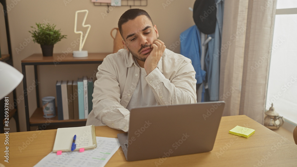 A pensive young hispanic man works from home on a laptop in a well-organized living room. A pensive young hispanic man works from home on a laptop in a well-organized living room.