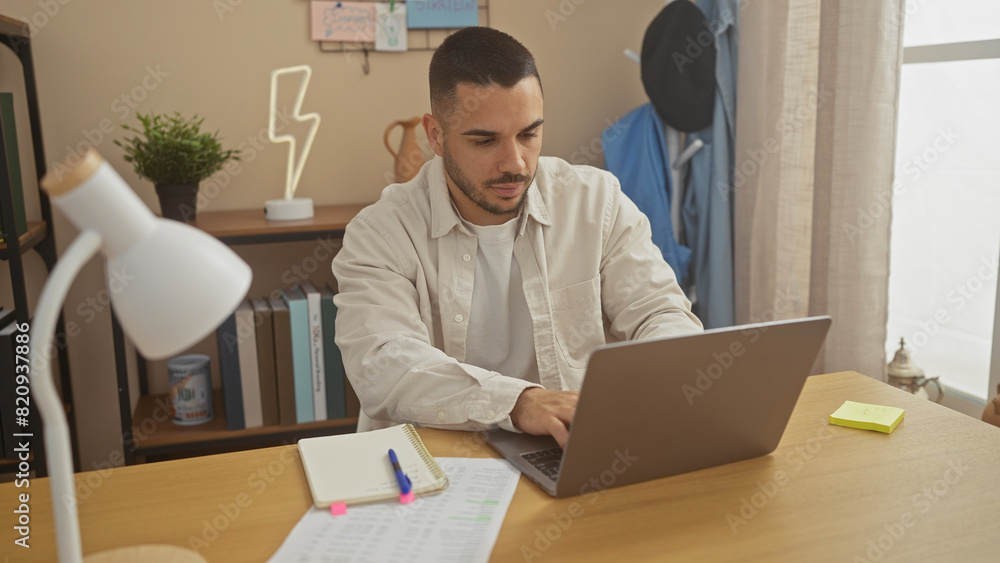 Hispanic man working from home using laptop at modern apartment's living room desk. Hispanic man working from home using laptop at modern apartment's living room desk.