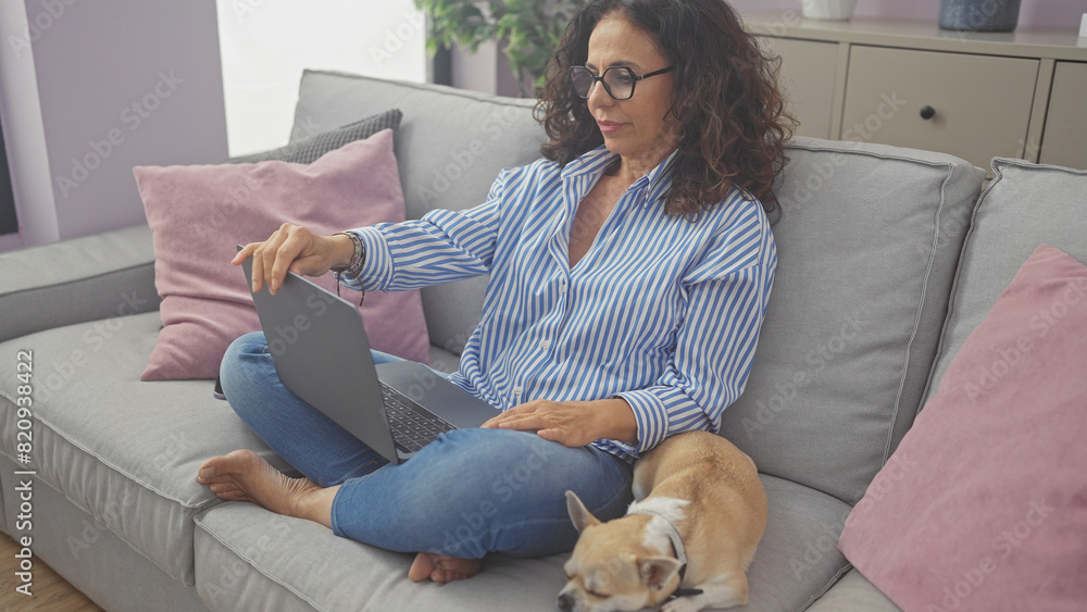 A middle-aged hispanic woman works on a laptop at home with her chihuahua resting beside her on the couch. A middle-aged hispanic woman works on a laptop at home with her chihuahua resting beside her on the couch.