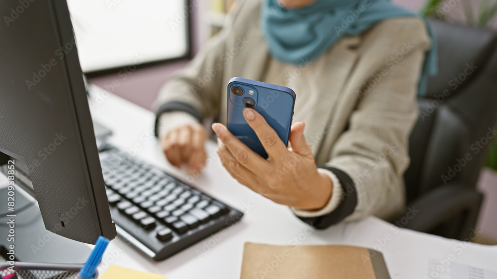 A mature hispanic woman multitasks with a smartphone and computer keyboard at her office desk. A mature hispanic woman multitasks with a smartphone and computer keyboard at her office desk.