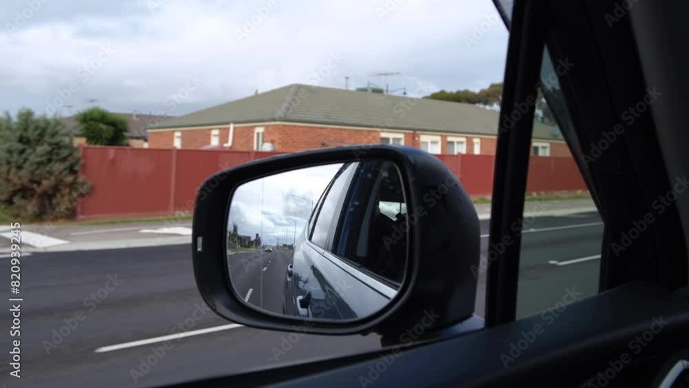 Close-up of a side-view mirror of a moving car with a blurry view of a row of suburban houses on the roadside. Concept of driving on a main road with multiple lanes in an Australian neighborhood. Close-up of a side-view mirror of a moving car with a blurry view of a row of suburban houses on the roadside. Concept of driving on a main road with multiple lanes in an Australian neighborhood.