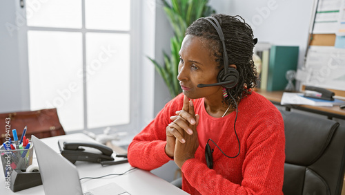 Fotografi A contemplative african american woman in a headset sits at her office desk, surrounded by modern technology