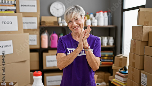 A smiling middle-aged woman with gray hair wearing a volunteer t-shirt claps in a donation center surrounded by boxes. photo