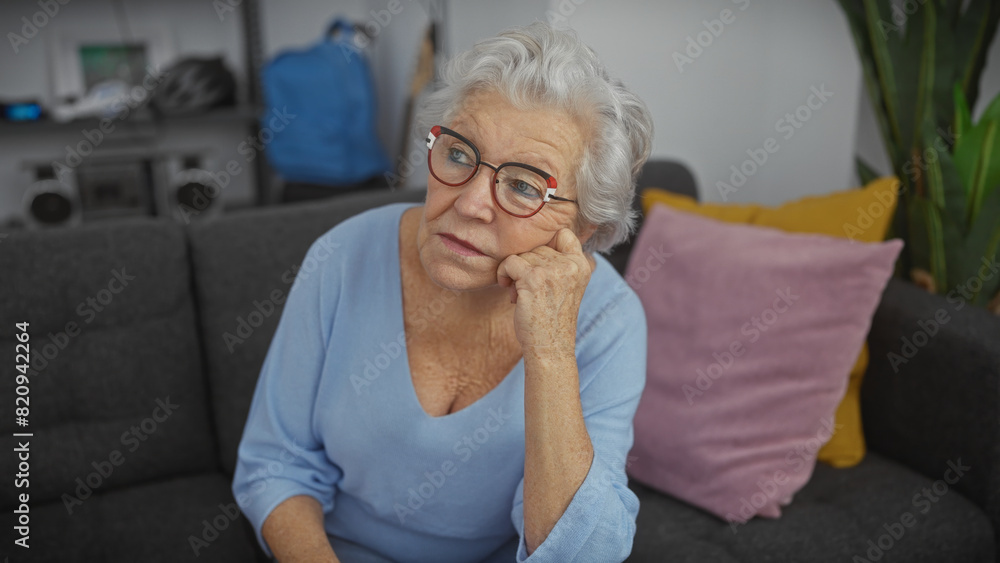 A thoughtful elderly woman with glasses sits on a sofa in a modern living room setting, expressing contemplation. A thoughtful elderly woman with glasses sits on a sofa in a modern living room setting, expressing contemplation.
