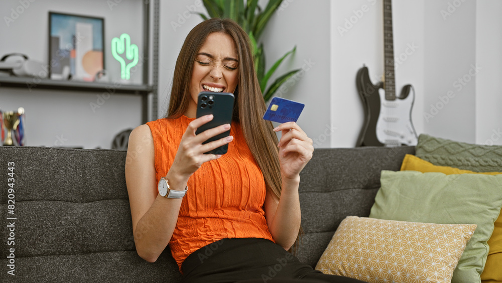 A cheerful woman using a smartphone and holding a credit card on a couch expressing online shopping joy in a modern living room. A cheerful woman using a smartphone and holding a credit card on a couch expressing online shopping joy in a modern living room.