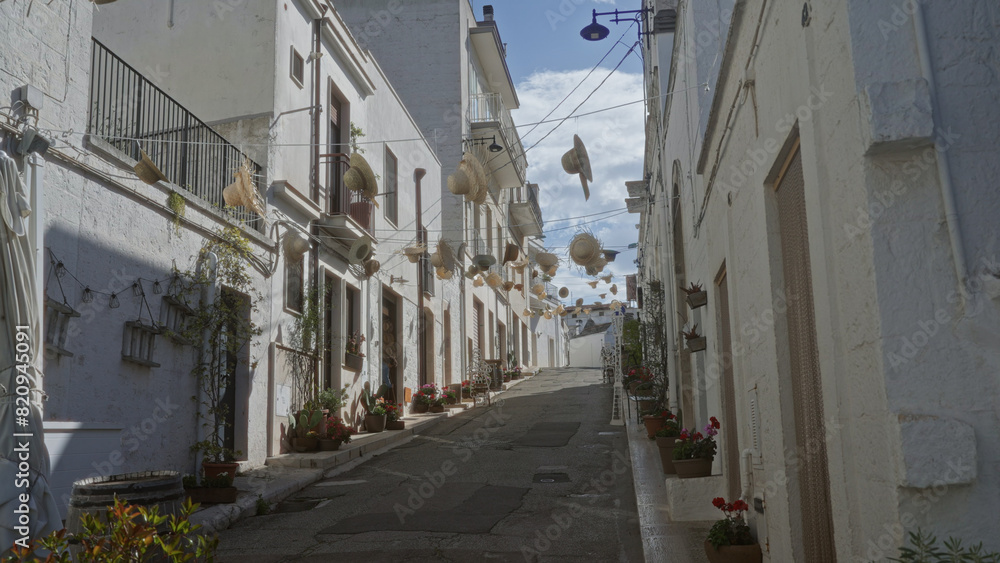 Charming street in alberobello, italy, puglia, decorated with hanging hats, flowers, and potted plants under a blue sky in a historic town setting. Charming street in alberobello, italy, puglia, decorated with hanging hats, flowers, and potted plants under a blue sky in a historic town setting.